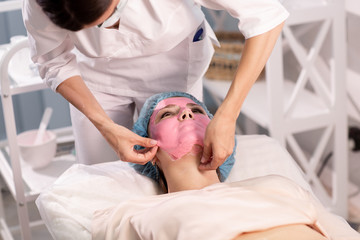 Fototapeta na wymiar The cosmetologist removes the pink wellness mask from the patient's face. Close up. Cosmetic procedures in the clinic