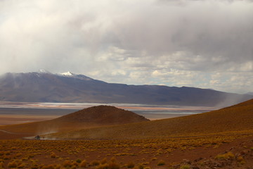 Fototapeta na wymiar Hills with brown soil in the foreground, a lake in the middle and mountains under a stormy sky in the background. Bolivia desert highlands, Potosi region 