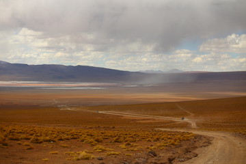 Valley with a dusty trail and  off road vehicles on it. Light brown soil and a lake far away. Mountains and stormy sky. Bolivia highlands desert. 