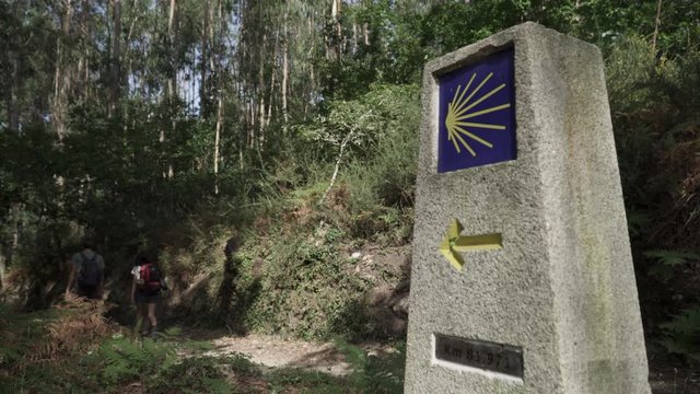 Way of St. James sign and pilgrims walking on a forest footpath. Yellow scallop sign pilgrimage to Santiago de Compostela