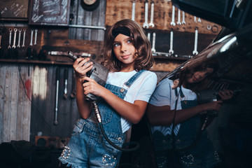 Little cute girl is posing with pneumatic drill for photographer at auto service workshop.