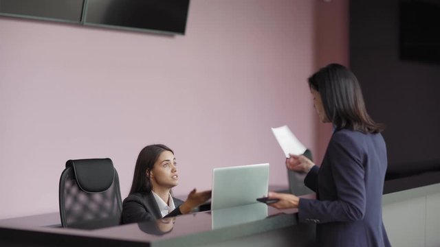 Middle aged businesswoman arguing with young receptionist at counter in business center. Female entrepreneur showing invitation letter, receptionist refusing to let her in