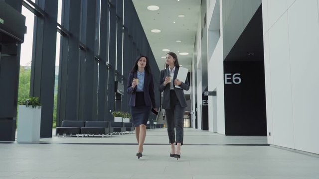 Full length slow motion shot of two businesswomen, young and middle aged, in formal suits walking down office hall together and talking. Colleagues holding takeaway coffee cups, cell phone and laptop