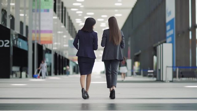 Rear view full length follow shot of two businesswomen in formal suits and high heels walking down office building lobby together in slow motion and talking
