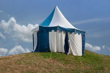 Blue and white medieval tent standing on the hill top with blue sky and white clouds in the background