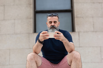 Bearded man sitting outside, in shorts and blue t-shirt, checking his phone