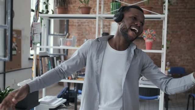 Happy african american man listening music from smart phone in wireless black headphones. Funny dancing and singing in the office. Smiling and laughing handsome young man indoors