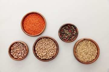 top view of wooden bowls with red lentil, beans, chickpea, oatmeal and peppercorns on white marble surface