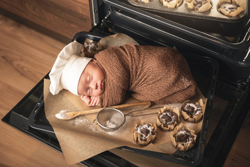 Newborn baby wearing chef's hat is lying on the oven tray with a muffins