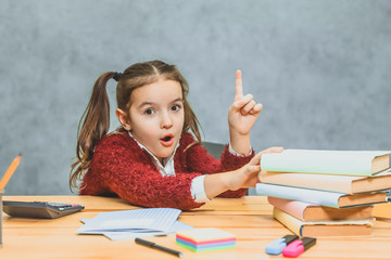 Very good schoolgirl girl sitting at the table. During this, he holds his hand on the books that stand on the table. Looking at the camera shows the index finger up. Has a surprised facial expression.