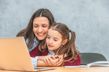 Mom and schoolgirl are sitting at the table. Mom hugs her daughter very much and looks at the laptop. Sincere emotions. The girl looks into a gray laptop.