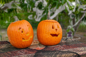 Two carved pumpkins sit in the foreground on a tablecloth covered table with spider web netting. The decorated background is out of focus to make the pumpkins stand out.