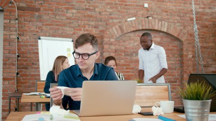 Ill Office Worker Sneezes, Suffering from Allergy, Flu or Cold at Loft Workplace with Laptop. Male Startup Employee Blows Nose in Paper Tissue at Work Desk. 4K Static Medium Indoors Shot