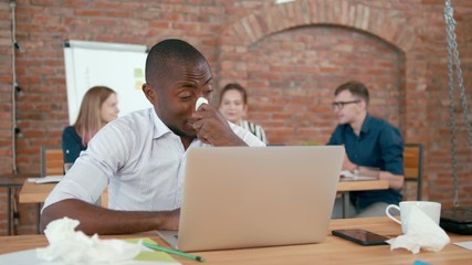 Ill African Office Worker Sneezes, Suffering from Allergy, Flu or Cold at Loft Workplace with Laptop. Male Startup Employee Blows Nose in Paper Tissue at Work Desk. 4K Static Medium Indoors Shot