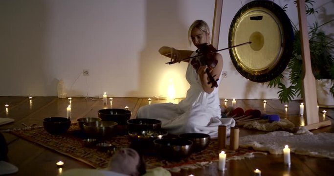 violinist woman is playing melody on violin during session of sound healing, people are lying on floor