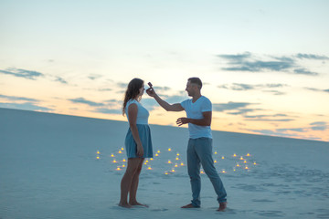 The guy makes the girl a marriage proposal by bending his knee while standing on the sand in the desert. Evening, candles burn in the sand.