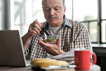 Senior hispanic man eating fresh salad in the morning sitting in the living room near laptop