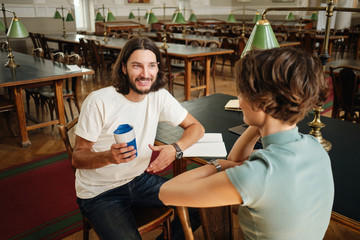 Young attractive male student happily talking with friend during study break in library of university