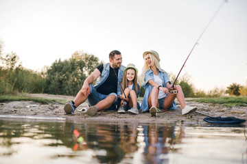 Fototapeta na wymiar Happy young family fishing on the lake. They smile, hold fishing rods and fishnets in their hands. The little daughter helps her parents fish. Wonderful landscape of the lake at sunset