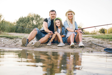 Fototapeta premium Happy young family fishing on the lake. They smile, hold fishing rods and fishnets in their hands. The little daughter helps her parents fish. Wonderful landscape of the lake at sunset
