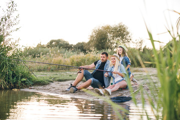 Happy young family fishing on the lake. They smile, hold fishing rods and fishnets in their hands. The little daughter helps her parents fish. Wonderful landscape of the lake at sunset