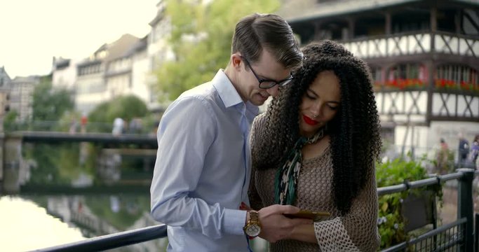young man is showing pictures on display of mobile phone to his girlfriend on bridge in small city