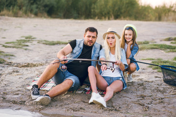 Fototapeta premium Happy young family fishing on the lake. They smile, hold fishing rods and fishnets in their hands. The little daughter helps her parents fish. Wonderful landscape of the lake at sunset