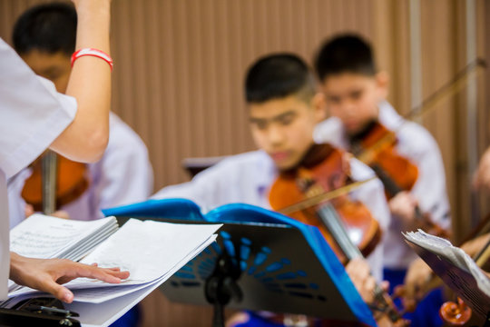 Selective Focus To Hands Of Conductor With Music Notation. Hands Of  Conductor With Blurry Asian Boy Students Playing Violin With Music Notation In The Group. Symphony Orchestra On Stage.