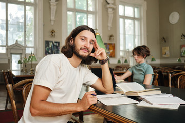 Young casual male student studying with book and conspectus while dreamily looking in camera in library of university