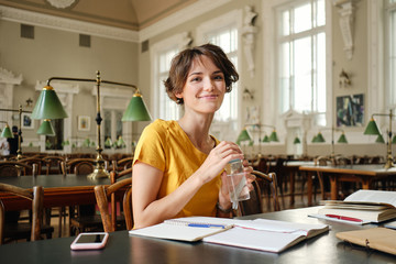 Young pretty smiling female student with water in hand joyfully looking in camera while study in library of university