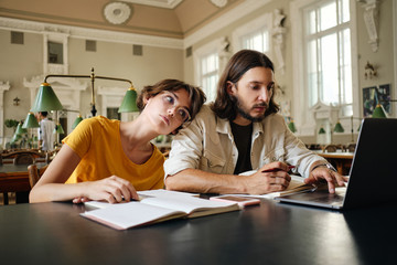 Two tired students thoughtfully working on study project with laptop and book in library of university