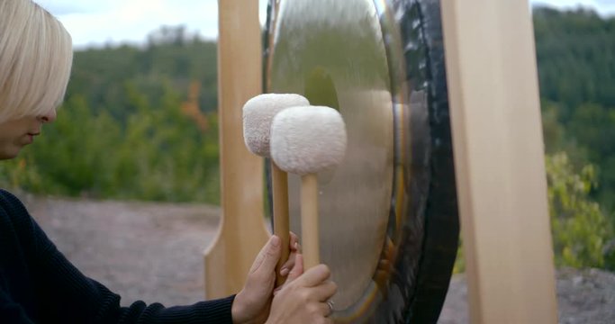 blonde woman is playing on gong, striking by two wooden mallets on nature
