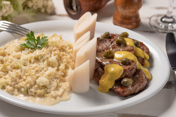 Risotto with filet mignon. Risoto and beef with capers and mustard in a white plate on wooden white background. Soft light. Italian food.