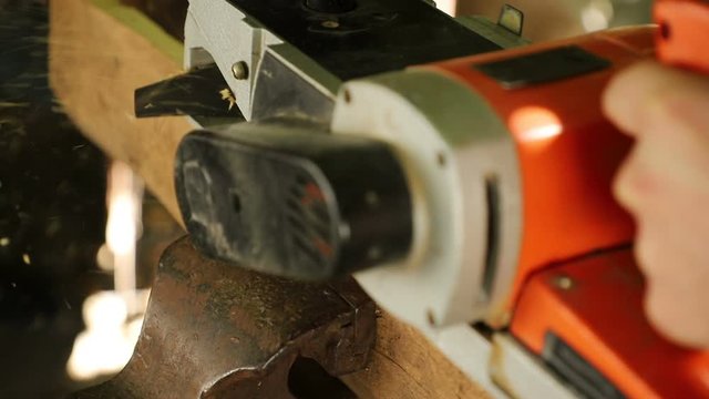 A carpenter handles the end of an old board with an electric planer, shavings scatter to the side, outdoors, close-up