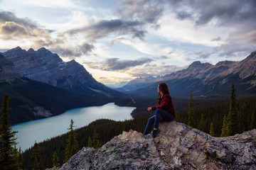 Naklejka premium Adventurous girl on the edge of a cliff overlooking the beautiful Canadian Rockies and Peyto Lake during a vibrant summer sunset. Taken in Banff National Park, Alberta, Canada.