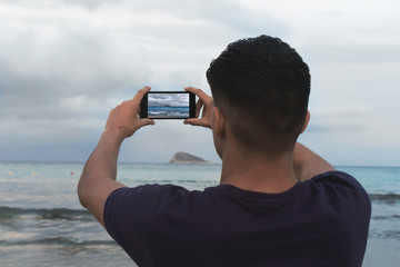 Brunette boy facing the sea, with very cloudy sky, taking a picture with a smartphone