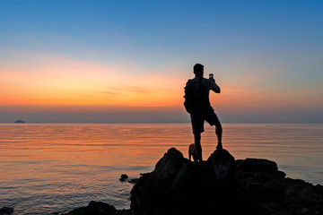 Silhouette of a dog and man with a backpack standing on the rock by the sea at sunset and taking pictures on a smartphone 
