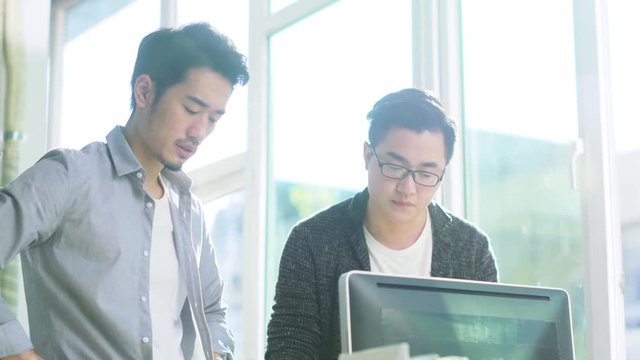 two young asian entrepreneurs standing by desk analyzing data using desktop computer