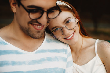 Obraz premium Close up portrait of a young couple sitting closely while smiling. Red haired girl with freckles looking at camera while leaning head on her boyfriend.