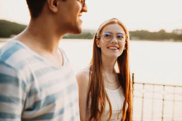 Young couple walking against sunset. Beautiful red haired woman with freckles looking at her boyfriend smiling.