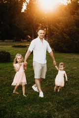 Outdoor shot of a young father walking hand in hand with his two daughters against sunset. Two little girls walking holding hand with their dad outdoor.