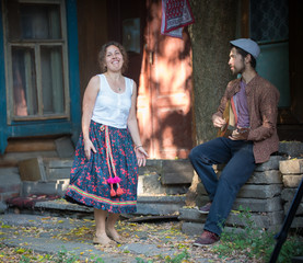 A smiling woman standing near the country house and a man playing balalaika