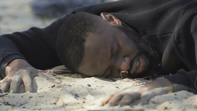 Afro-American male refugee suffering from thirst, lying on sand, lost in desert