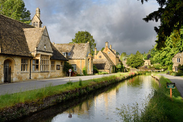 Morning sun on yellow Cotswold limestone buildings of Lower Slaughter on the River Eye with dark clouds Gloucestershire England