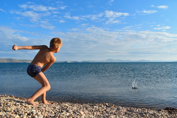 Fototapeta na wymiar Boy in swimming trunks on lake throws stones into water. Teenager plays on seashore. Summer vacation with parents on lake on fresh air.