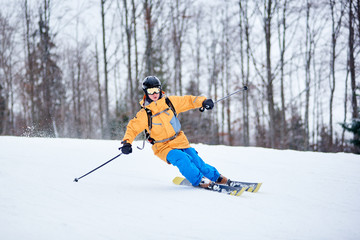 Front view of young man skier making turn on other side for next arc of skiing on snow-covered way. Professional skiing technique. Monochrome winter nature view. Bare trees on blurred background.