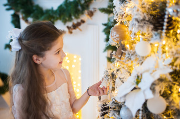 Beautiful little girl in elegant dress decorates a Christmas tree. Golden garland in the background