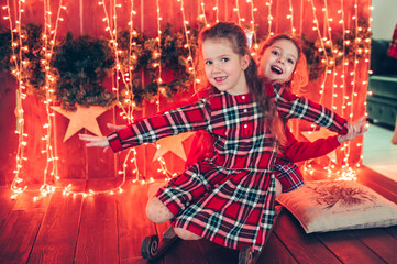 Two cute girls in red dresses are sitting on a wooden sled. Red background with golden garlands and bokeh.