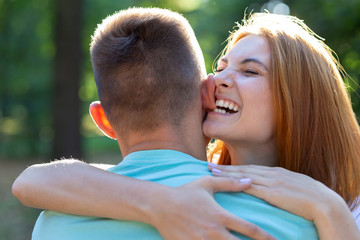 Beautiful teenage girl with bright red hair playfully biting her boyfriend by ear outdoors. First love and romantic relationship concept.