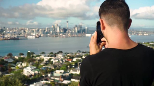 Young caucasian tourist taking a photo with phone of Auckland and Sky Tower from Mount Victoria, New Zealand